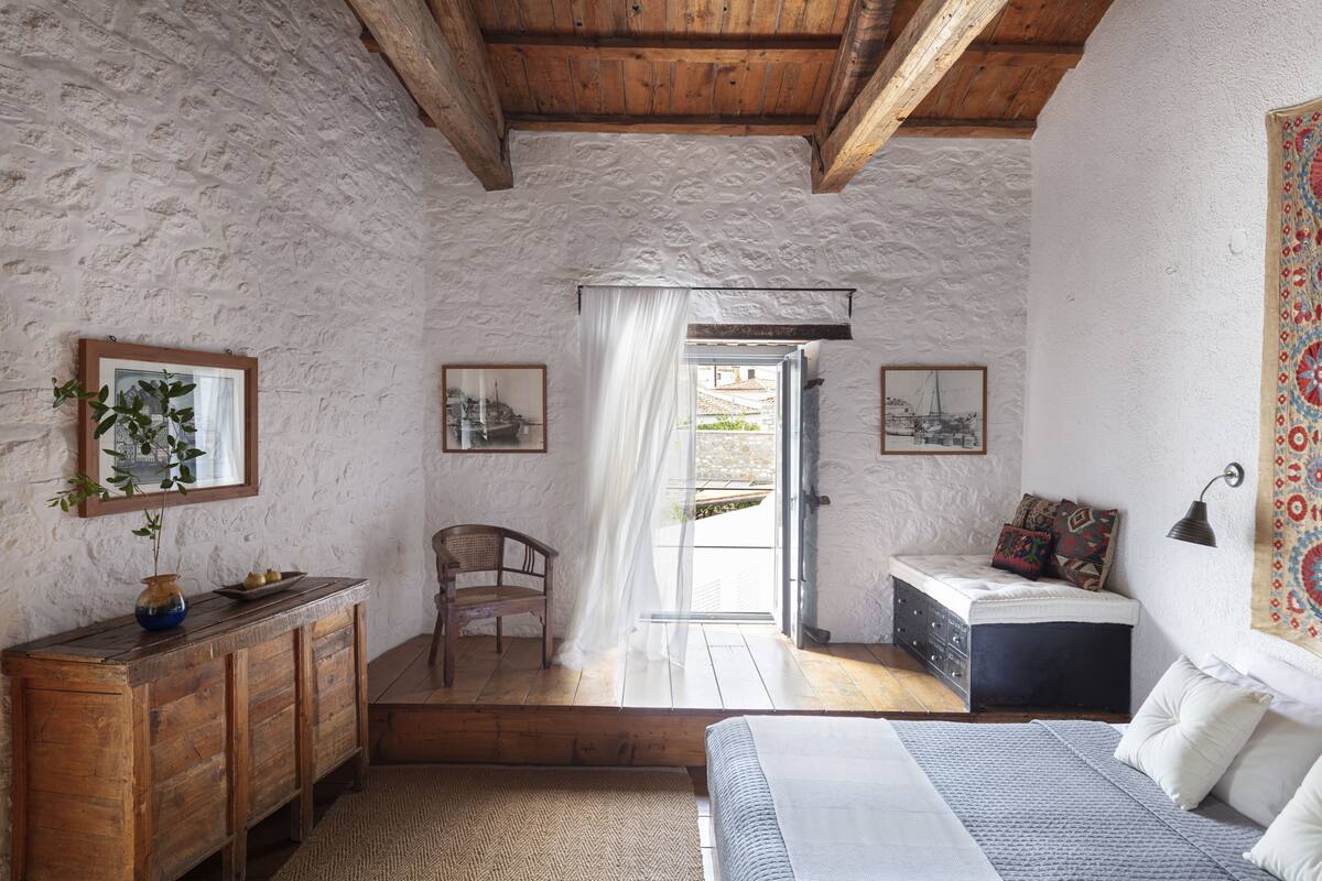 Une chambre à coucher avec un lit, une commode en bois, une chaise et un banc. Il y a une fenêtre donnant sur l’extérieur.