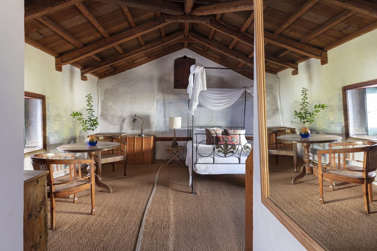 Une pièce rustique avec un haut plafond en bois, un lit à baldaquin blanc, des meubles en bois et une petite table sur laquelle est posé un vase.