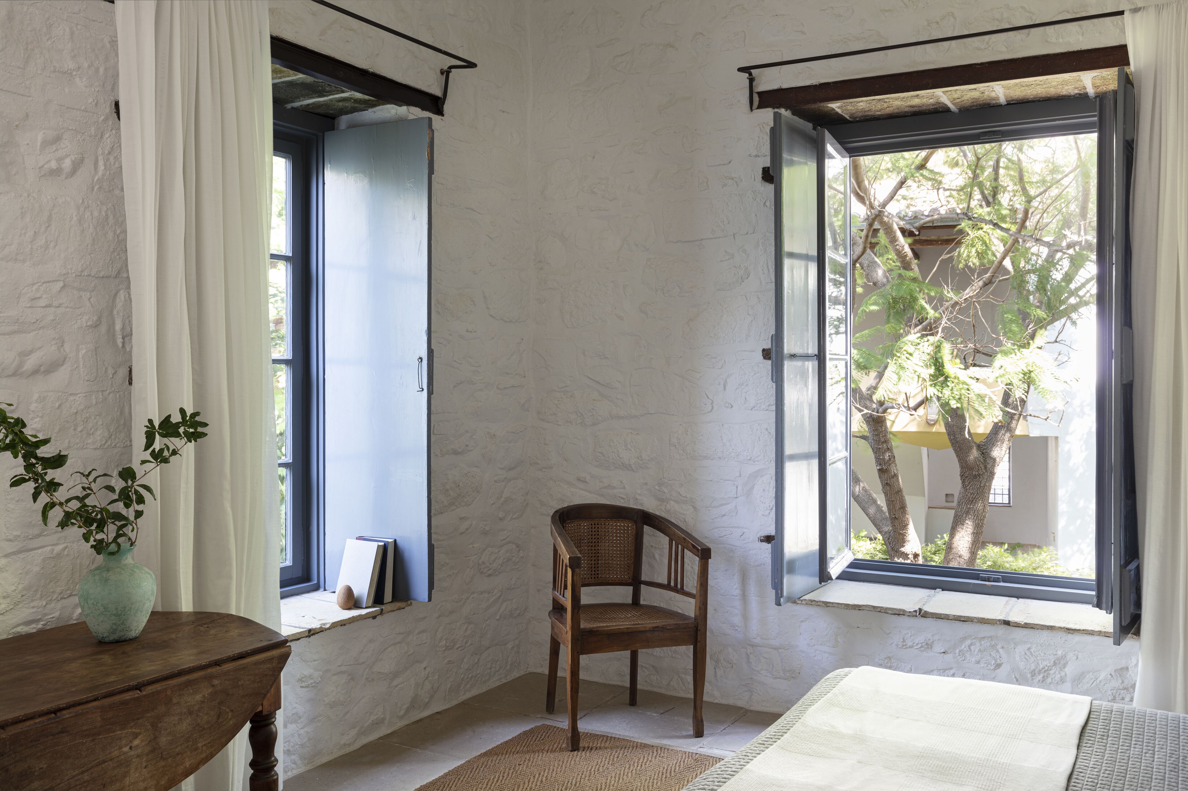 Una habitación con una silla de madera, una mesita con una planta y un ventanal con vistas a árboles y un edificio.