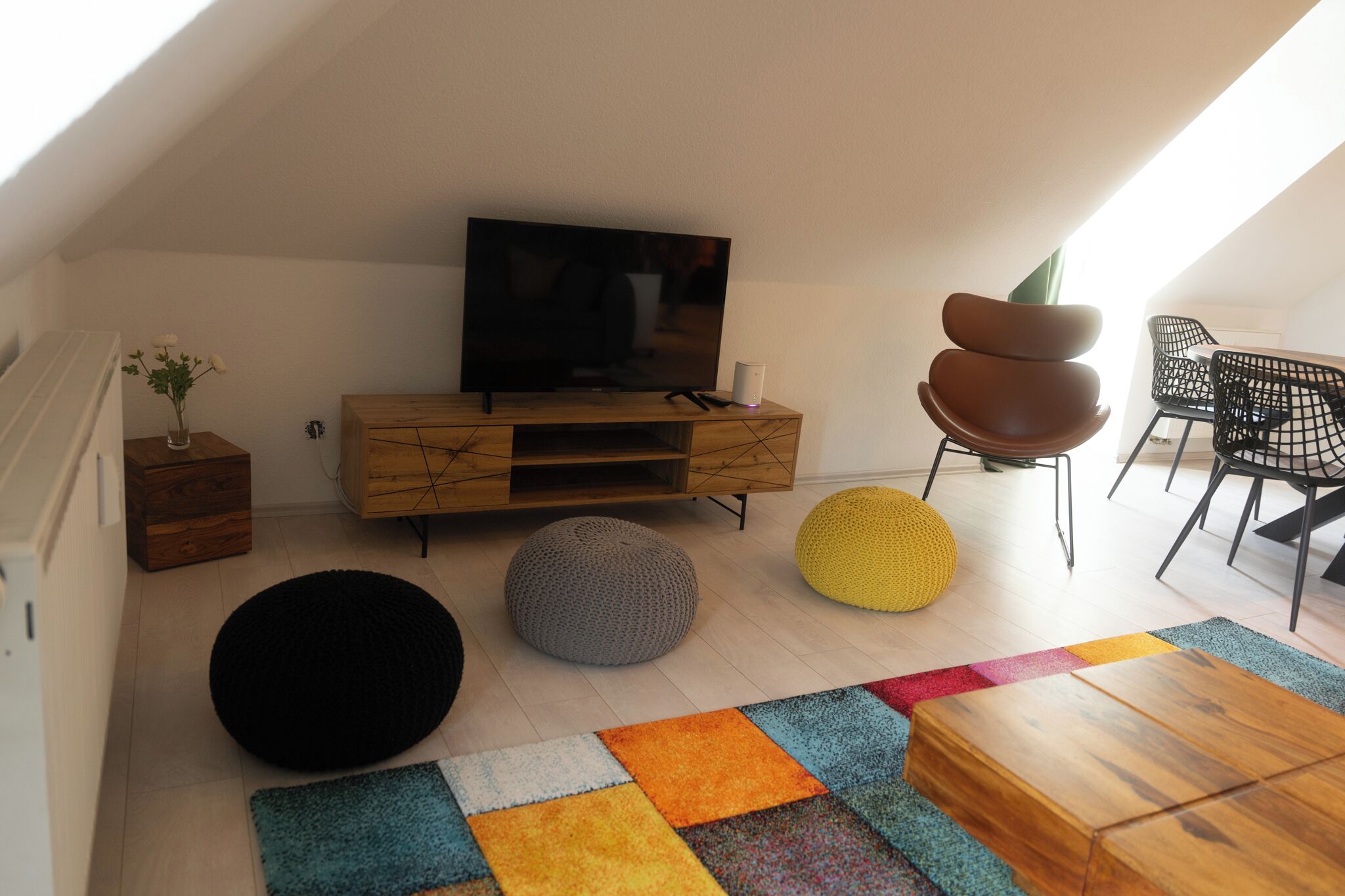 Ein modernes Wohnzimmer mit einem Fernseher auf einer Holzkommode, zwei Sitzsäcken, einem Couchtisch und einem Essbereich mit Stühlen.