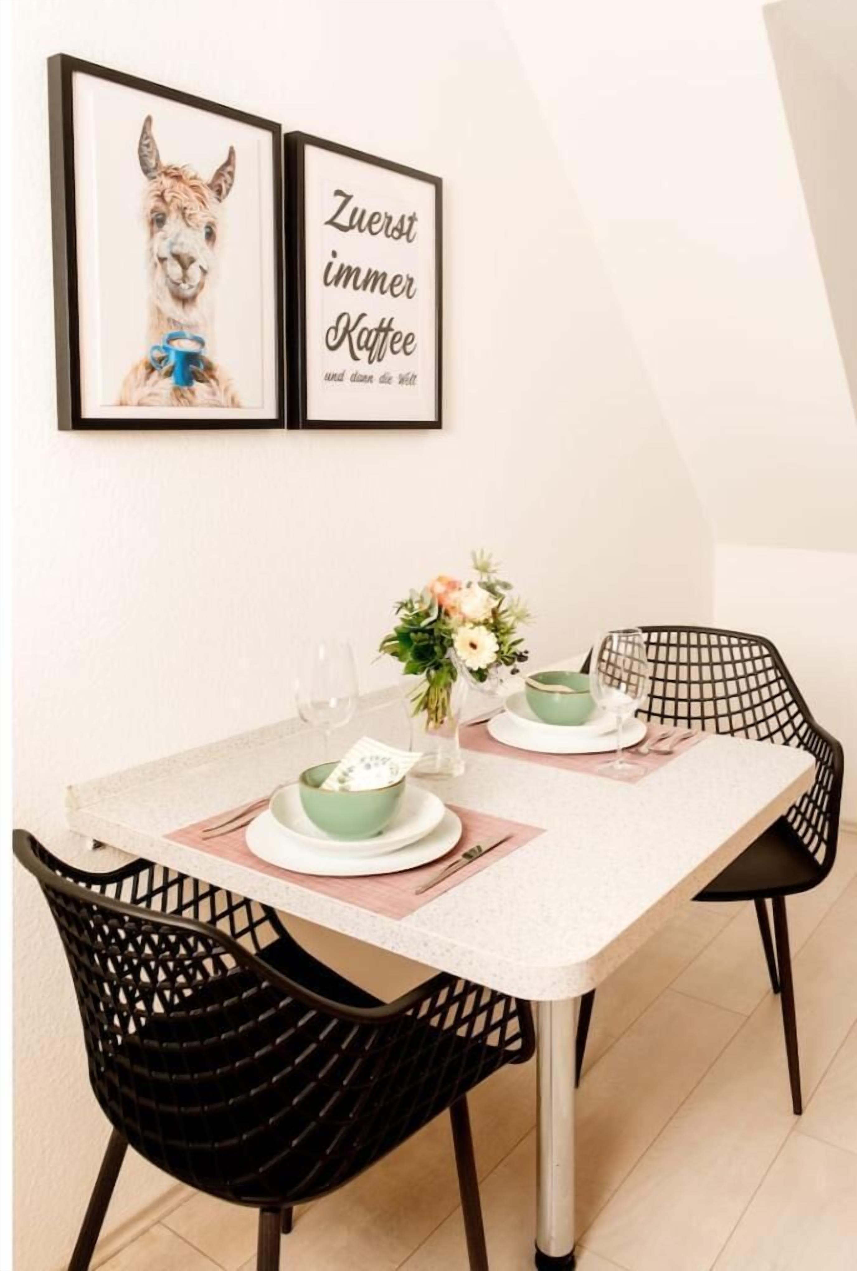 Een eethoek met een witte tafel gedekt voor twee personen, zwarte stoelen, een afbeelding van een lama in een lijst en een citaat in een lijst.
