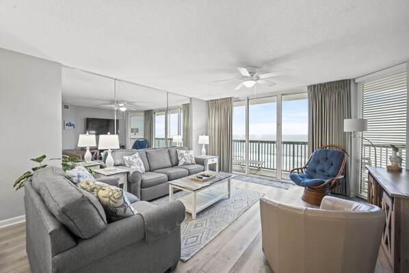 A modern living room with a grey sofa, a blue armchair, and a wooden coffee table. There is a large window with a view of the ocean.
