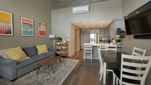 Una sala de estar con sofá, mesa de centro y una obra de arte en la pared. Hay una zona de cocina con mesa y sillas.
