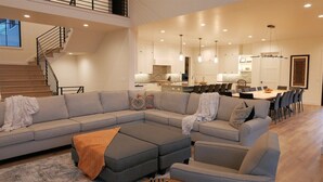 A modern living room with a sectional sofa, ottoman, and a dining area with a large table and chairs. - The Pickleball Gathering Place (Huntsville)