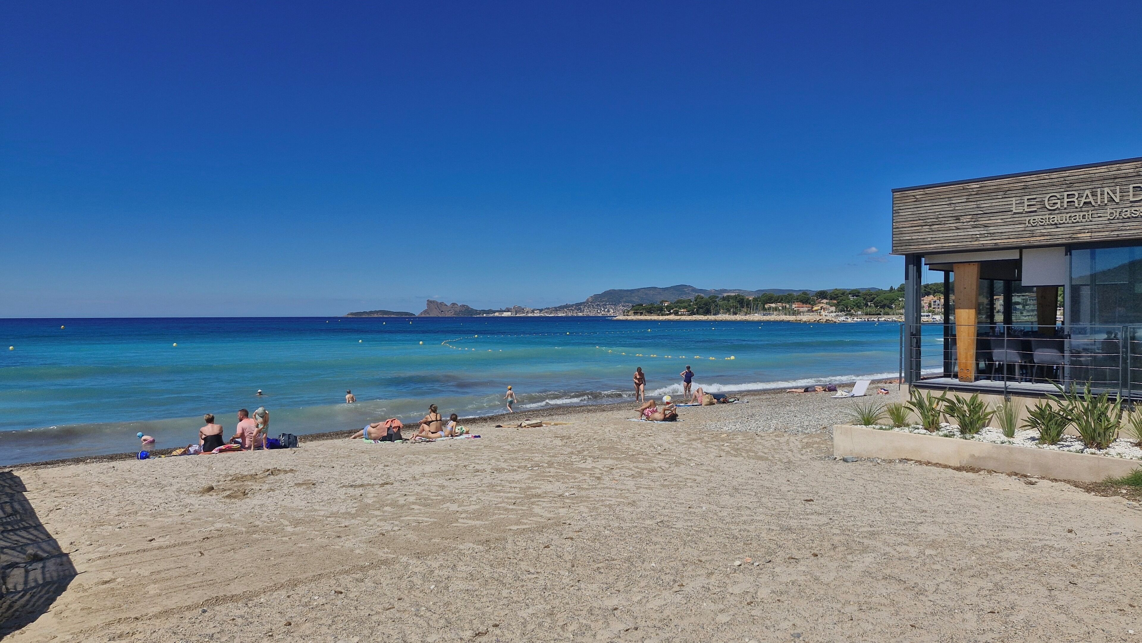 A beach with people sunbathing and swimming, a restaurant named "LE GRAIN" with outdoor seating, and a clear view of the sea and distant hills.