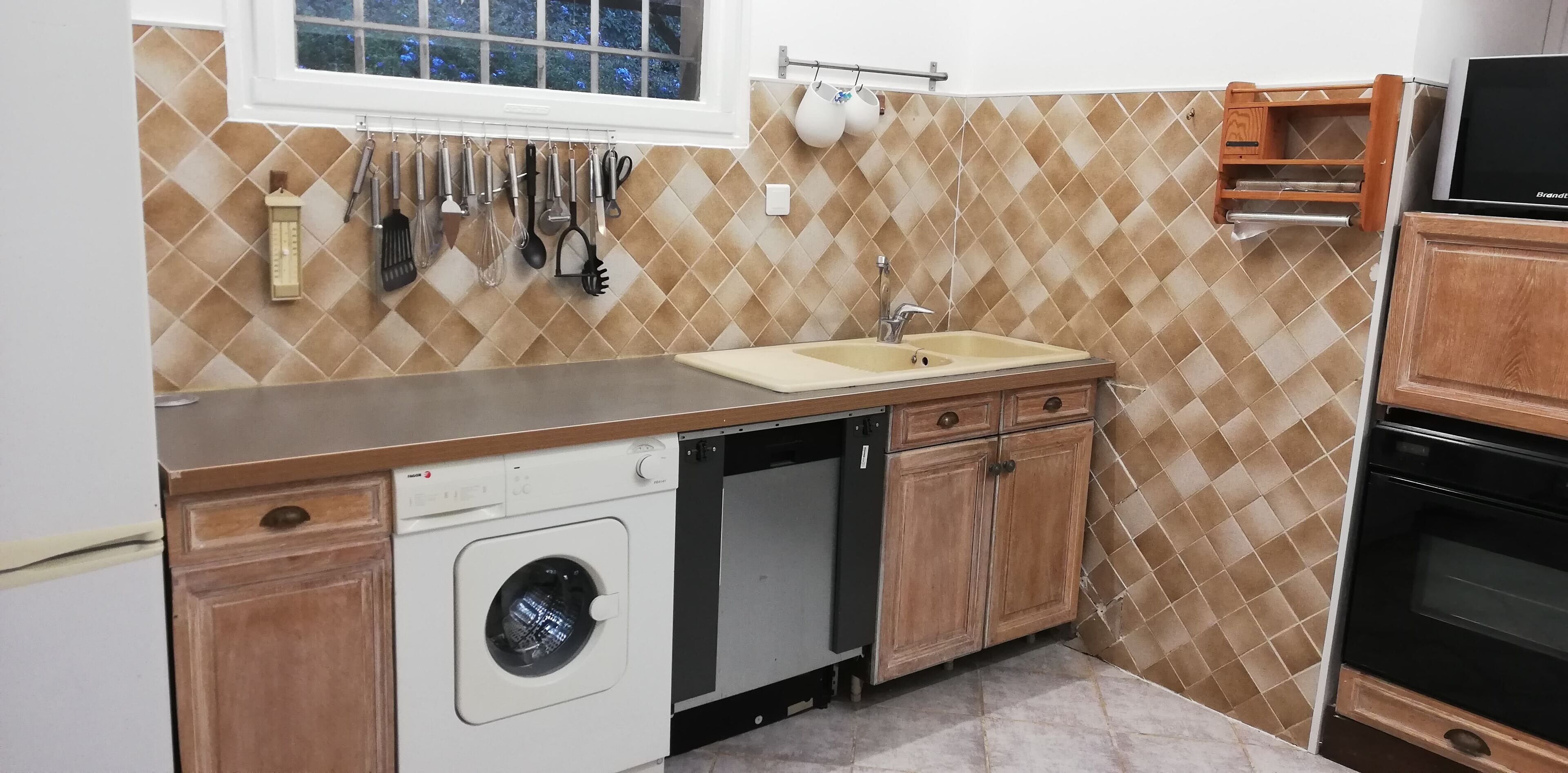 A kitchen with a dishwasher, sink, wooden cabinets, and a window with hanging utensils.