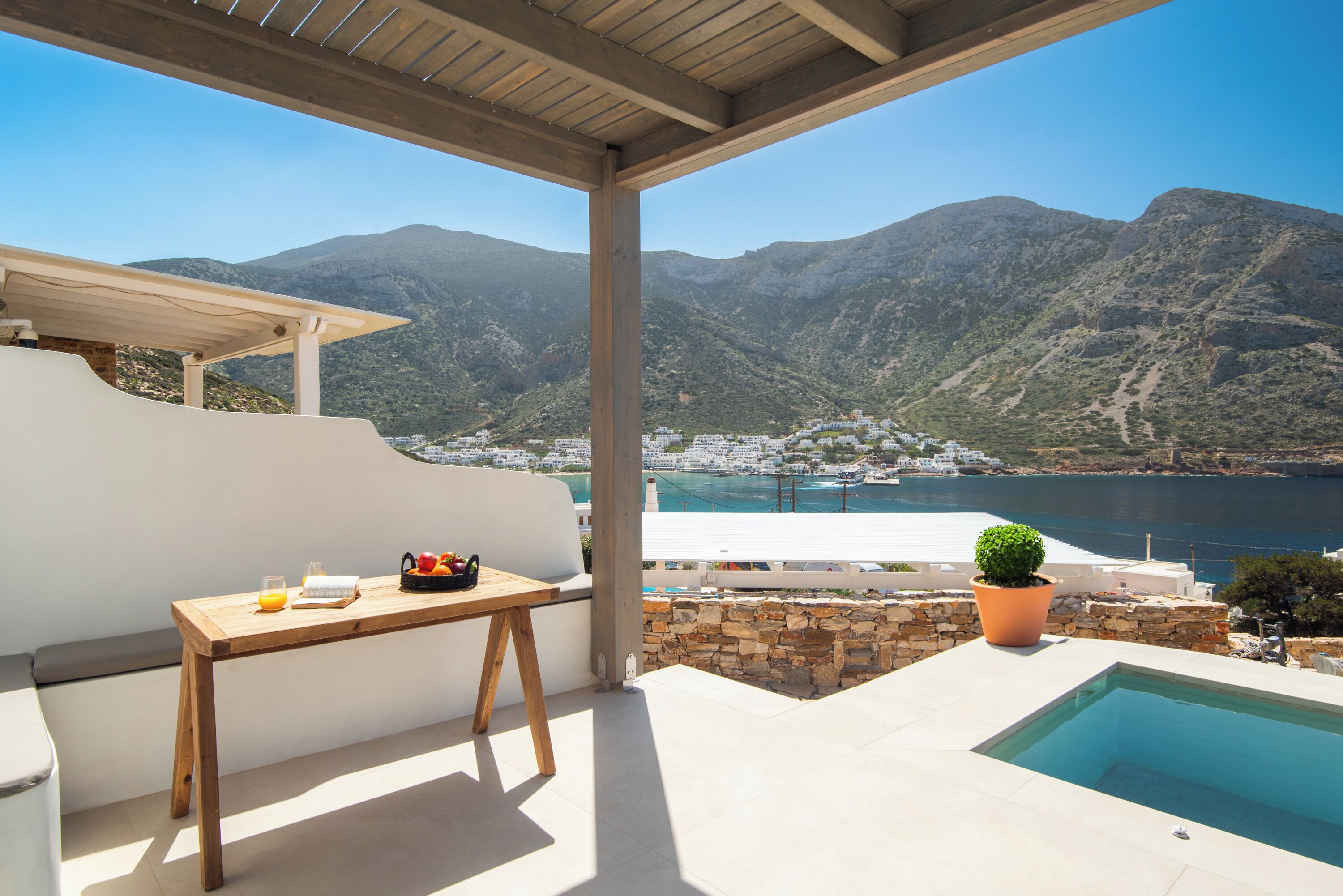 Una terraza con una mesa de madera, una planta en maceta y vistas a un paisaje montañoso y un cuerpo de agua.