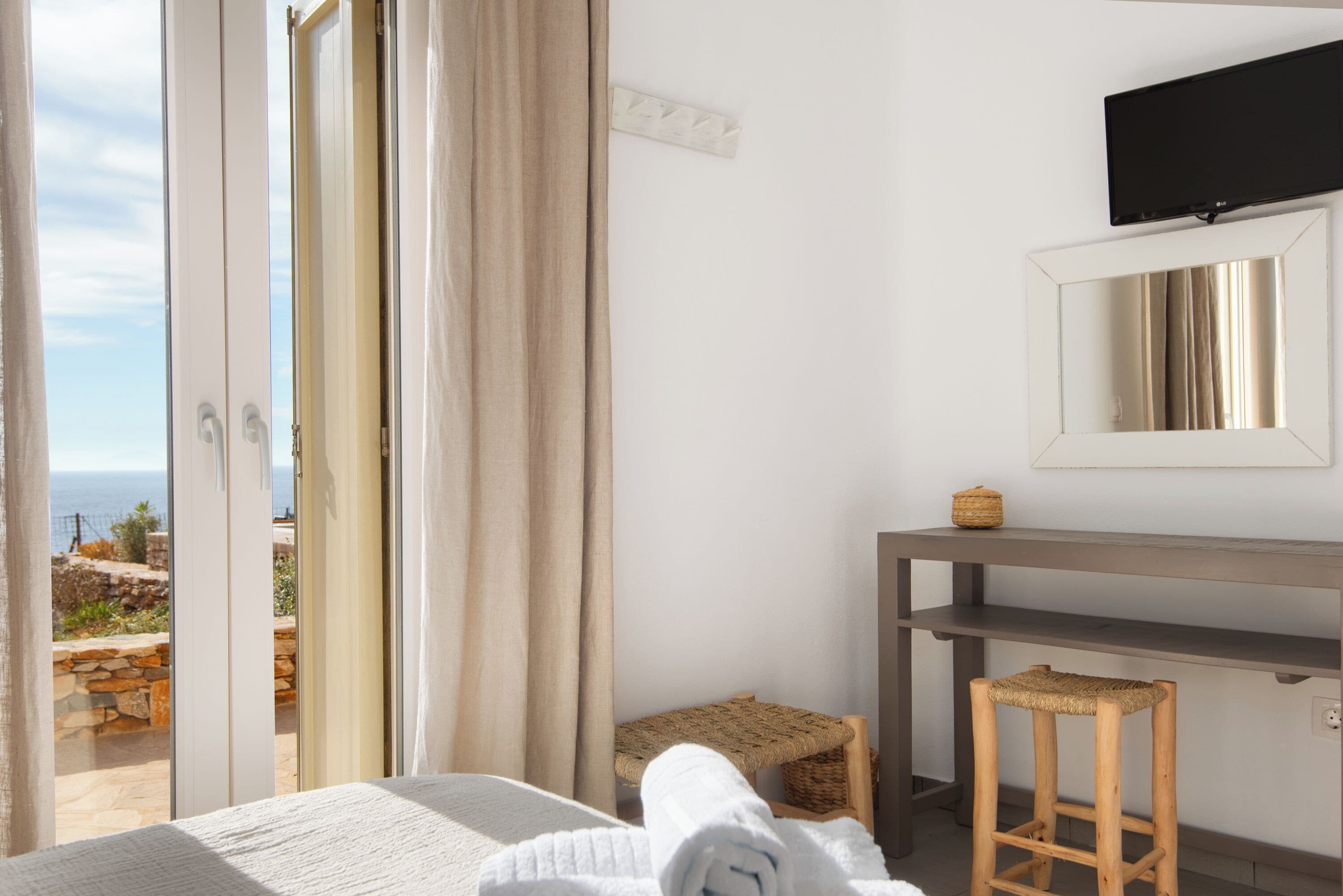Ein Zimmer mit einem Bett, einem kleinen Hocker, einem Tisch, einem Fernseher und Blick ins Freie.