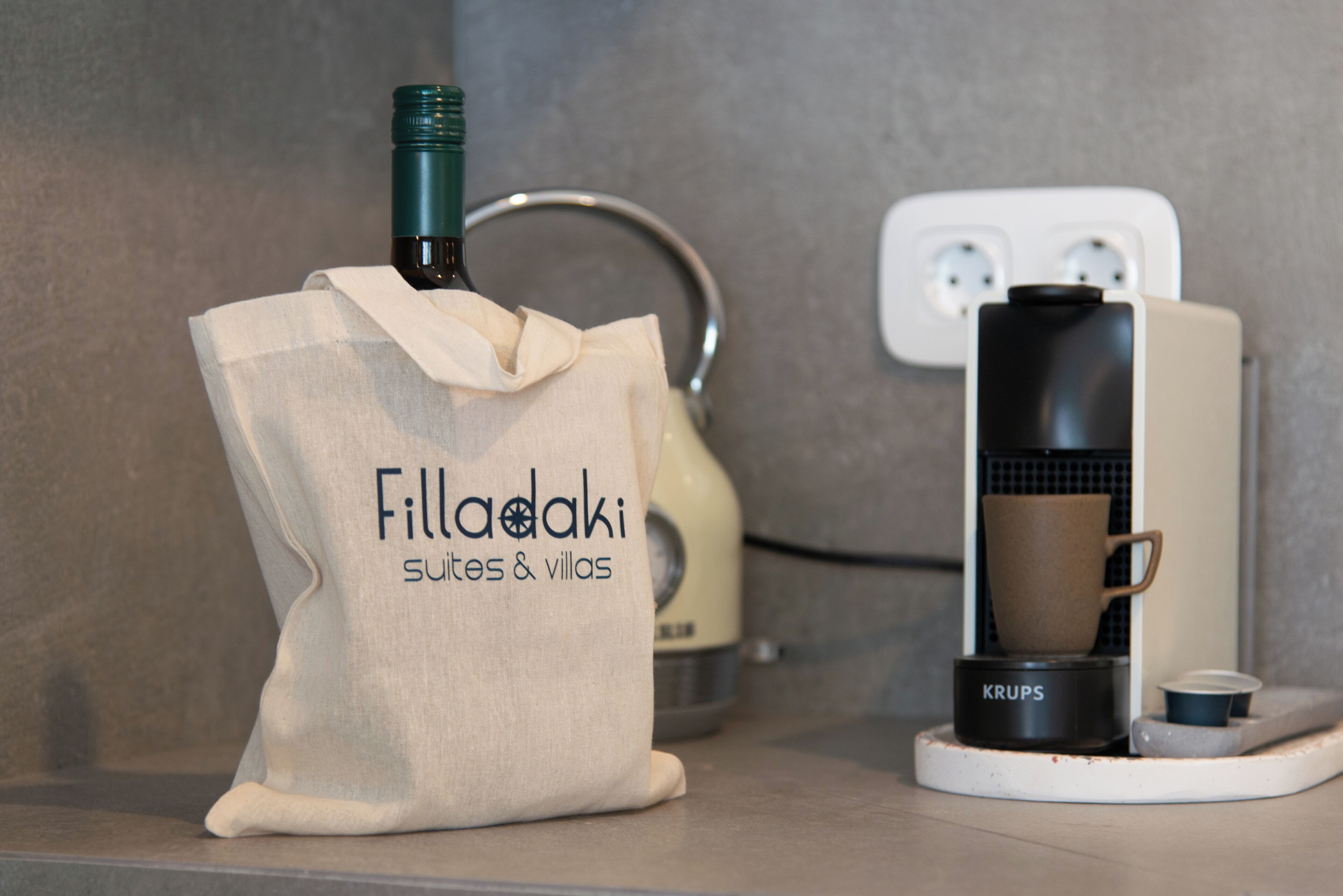 Un coin cuisine équipé d’une machine à café, d’un sac étiqueté « Filladaki suites & villas » et d’une prise de courant.
