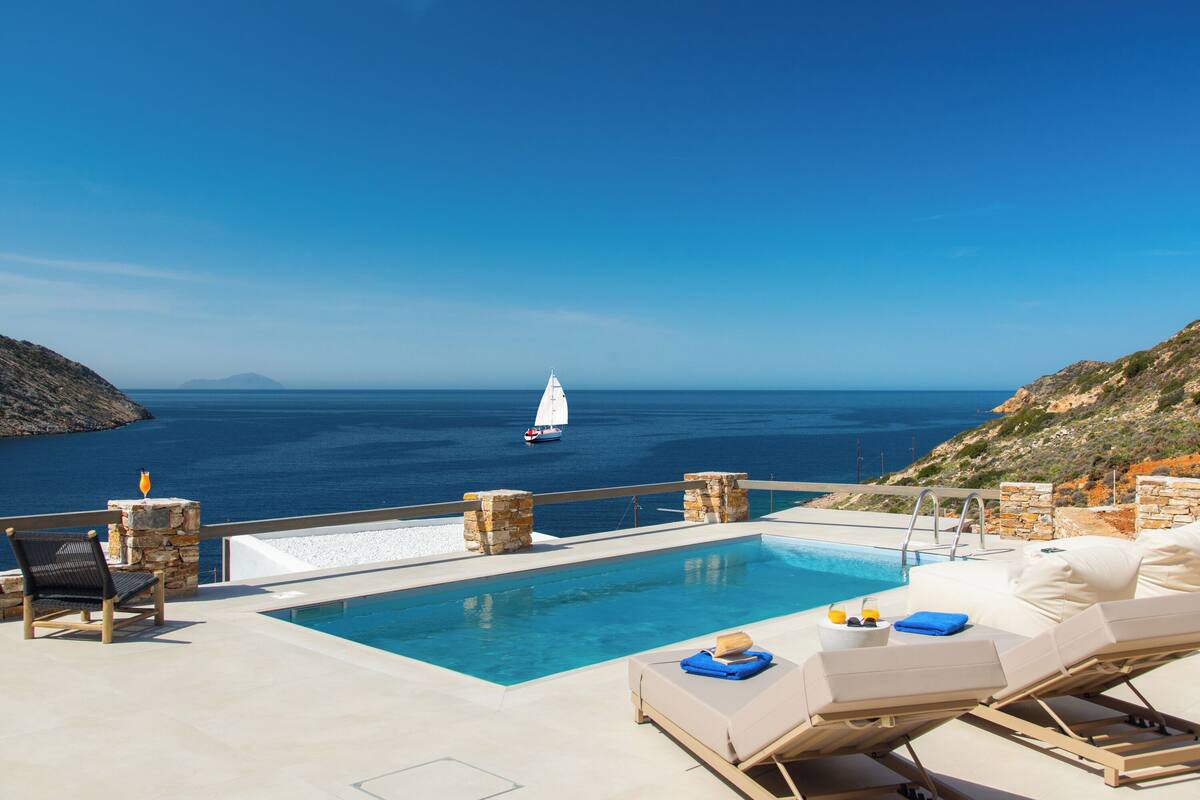 A poolside area with a view of the sea, a sailboat, and stone pillars.