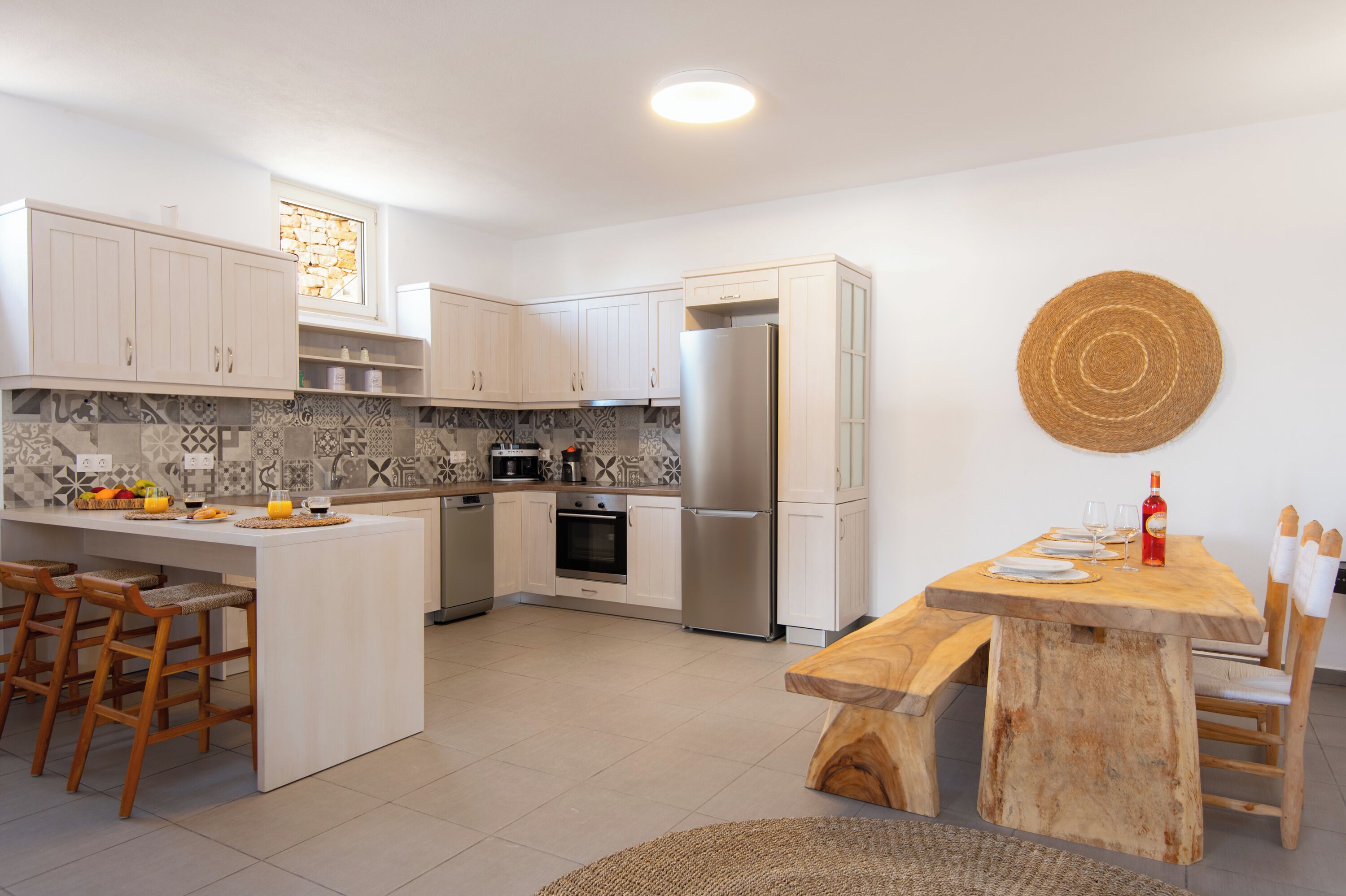 Una cocina moderna con armarios blancos, horno empotrado y refrigerador de acero inoxidable. Hay una mesa de comedor de madera con sillas y un mantel tejido.