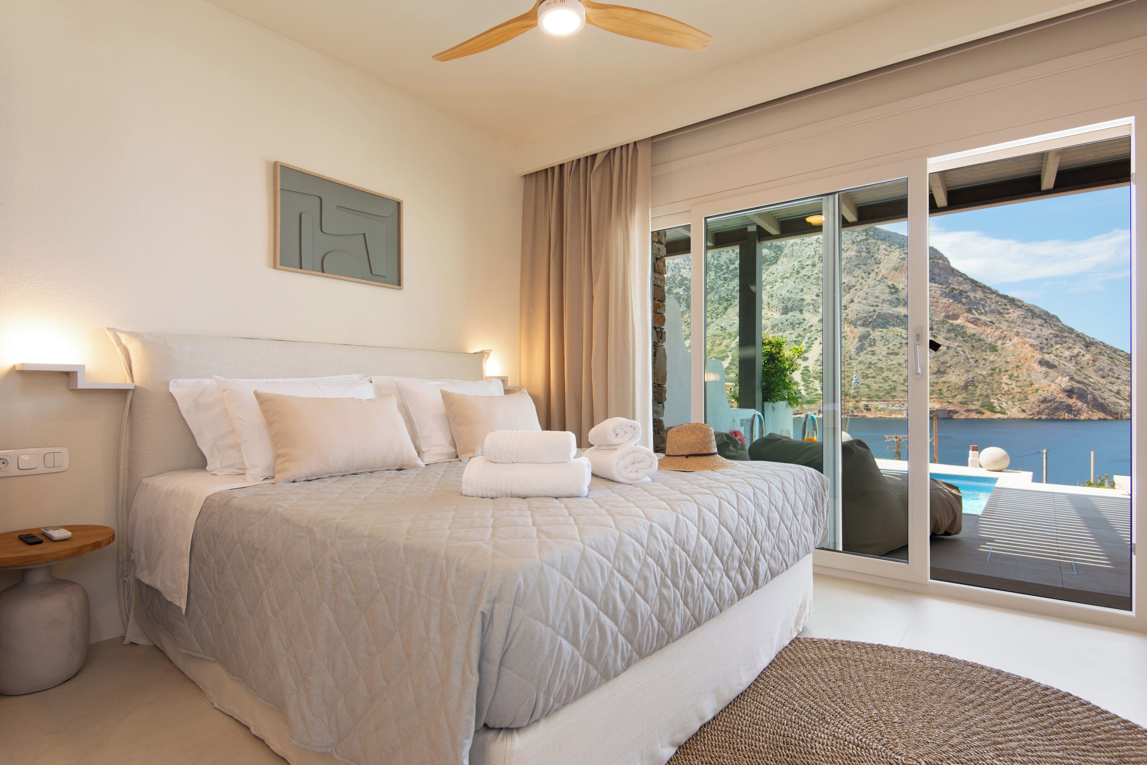 A bedroom with a large bed, a ceiling fan, a nightstand, and a view of a pool and mountains through a sliding glass door.