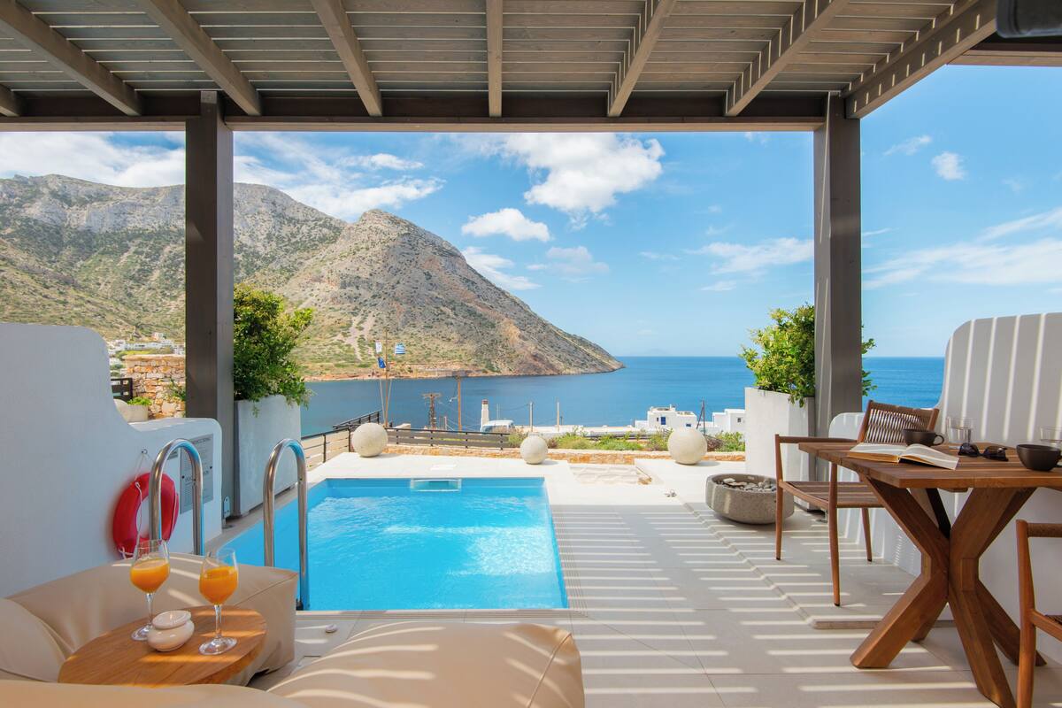 A pool area with a view of the sea and mountains, a wooden table with a book and glasses, and a lounge chair.