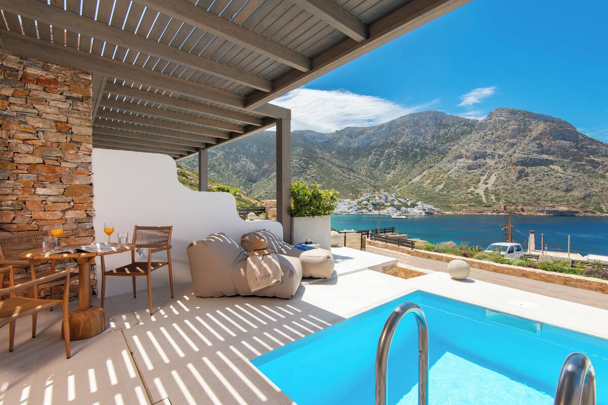 A pool area with a view of the sea and mountains, wooden furniture, and a stone wall.
