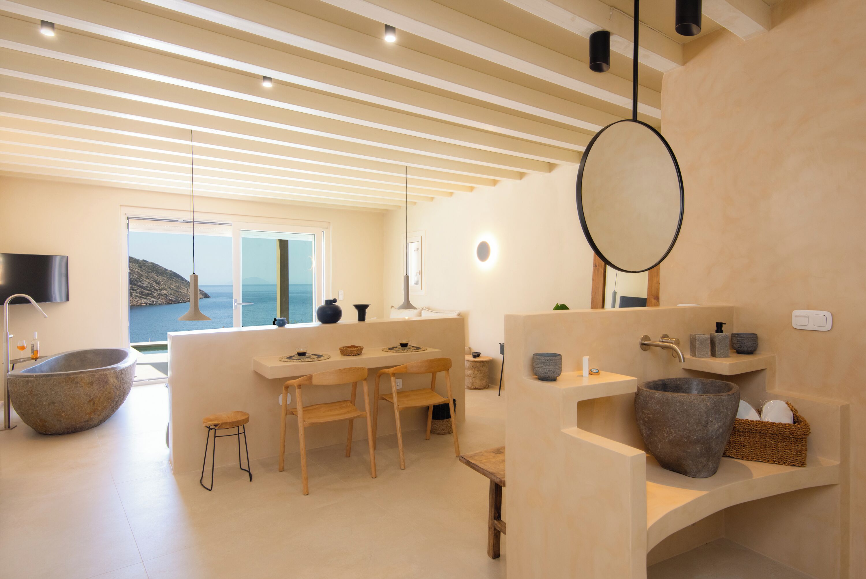 A modern bathroom with a large stone basin, wooden chairs, and a view of the sea.