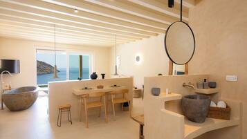 A modern bathroom with a large stone basin, wooden chairs, and a view of the sea.