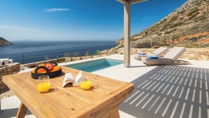 A wooden table with a fruit basket and drinks, a poolside lounge area, and a view of the sea. - Filadaki Villas (Sifnos)