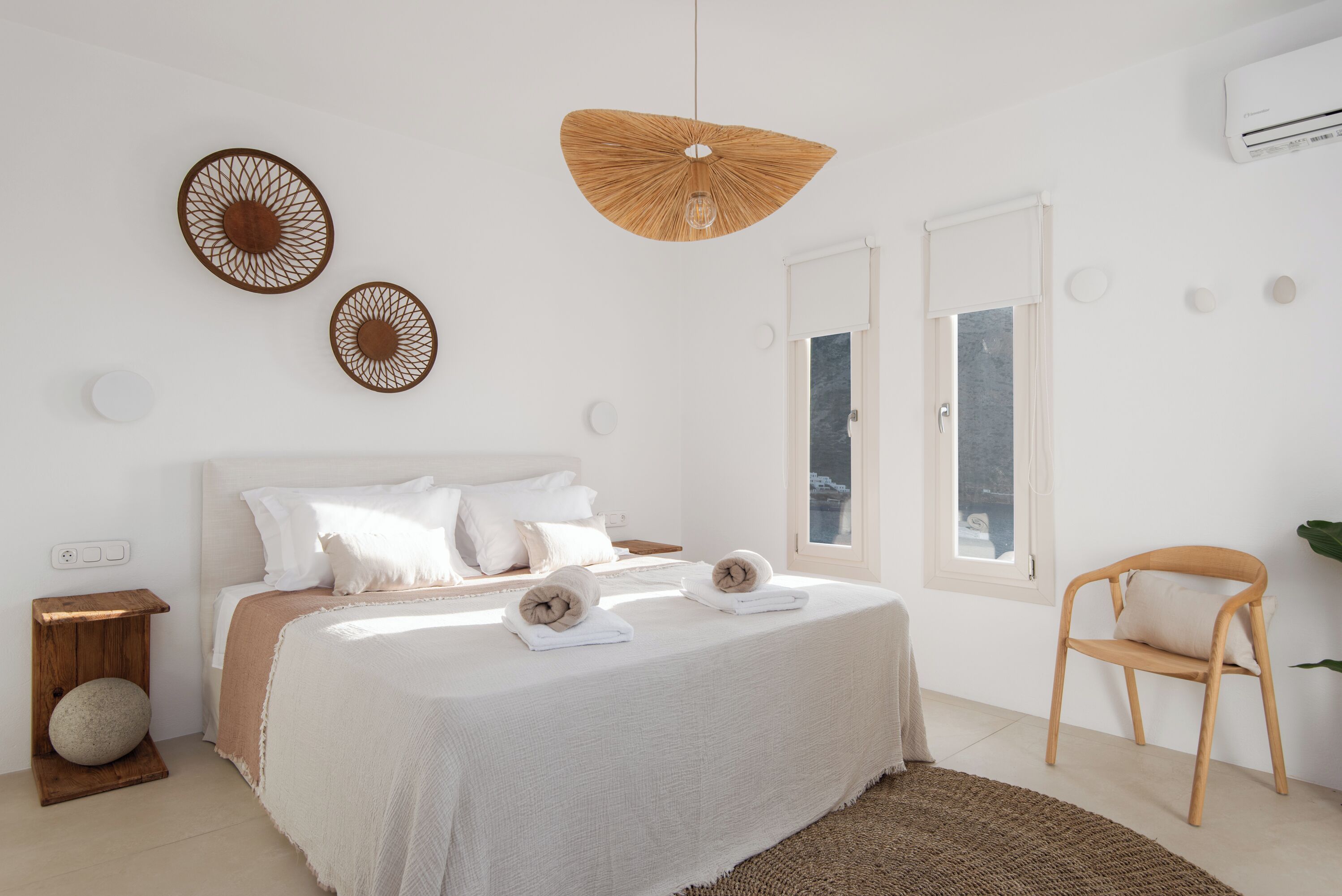 A minimalist bedroom with a large bed, two wall-mounted decorative items, a hanging light fixture, and a window with a view.