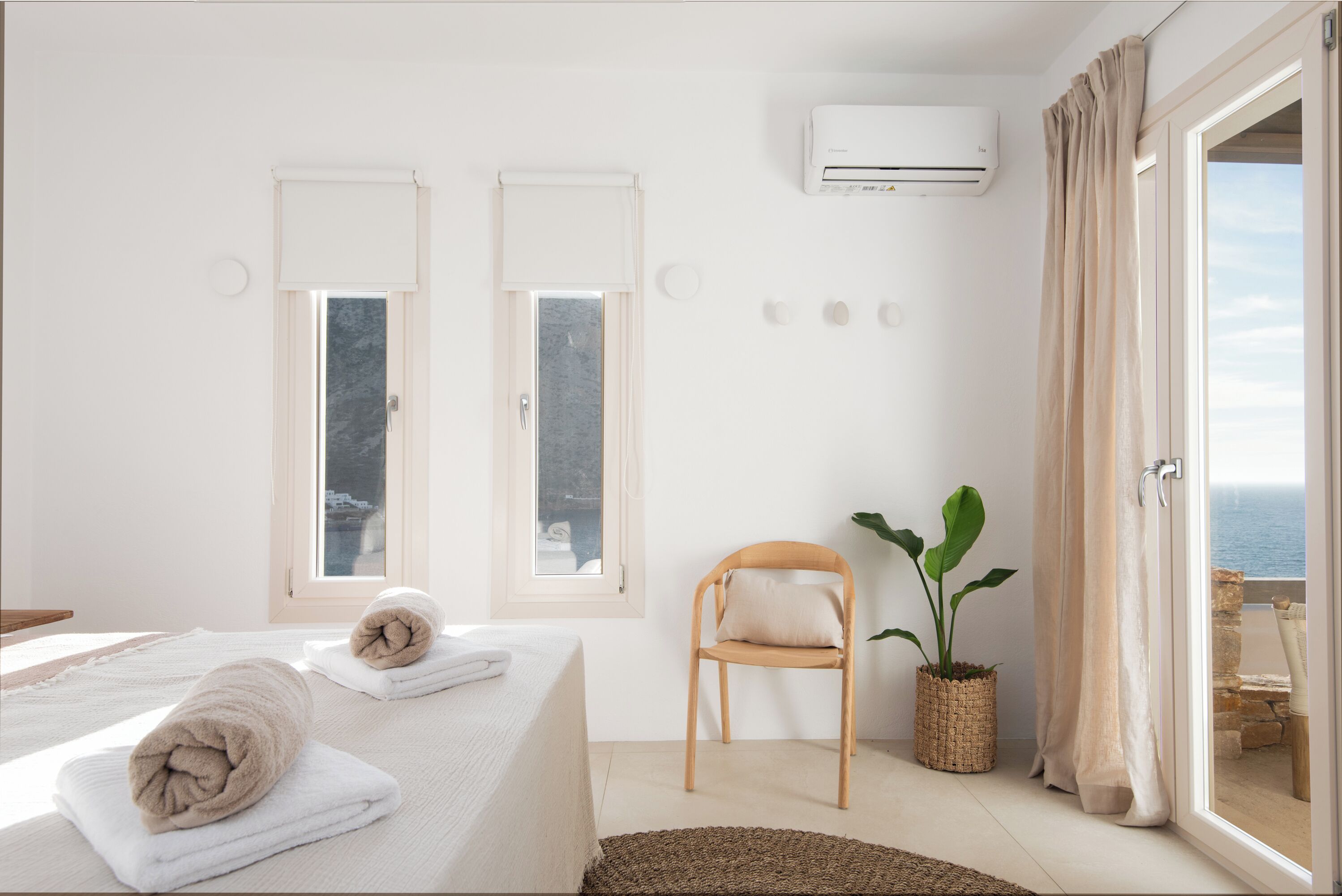 Quarto com cama, duas janelas, uma cadeira, planta em vaso e vista para o mar.