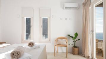 Quarto com cama, duas janelas, uma cadeira, planta em vaso e vista para o mar.