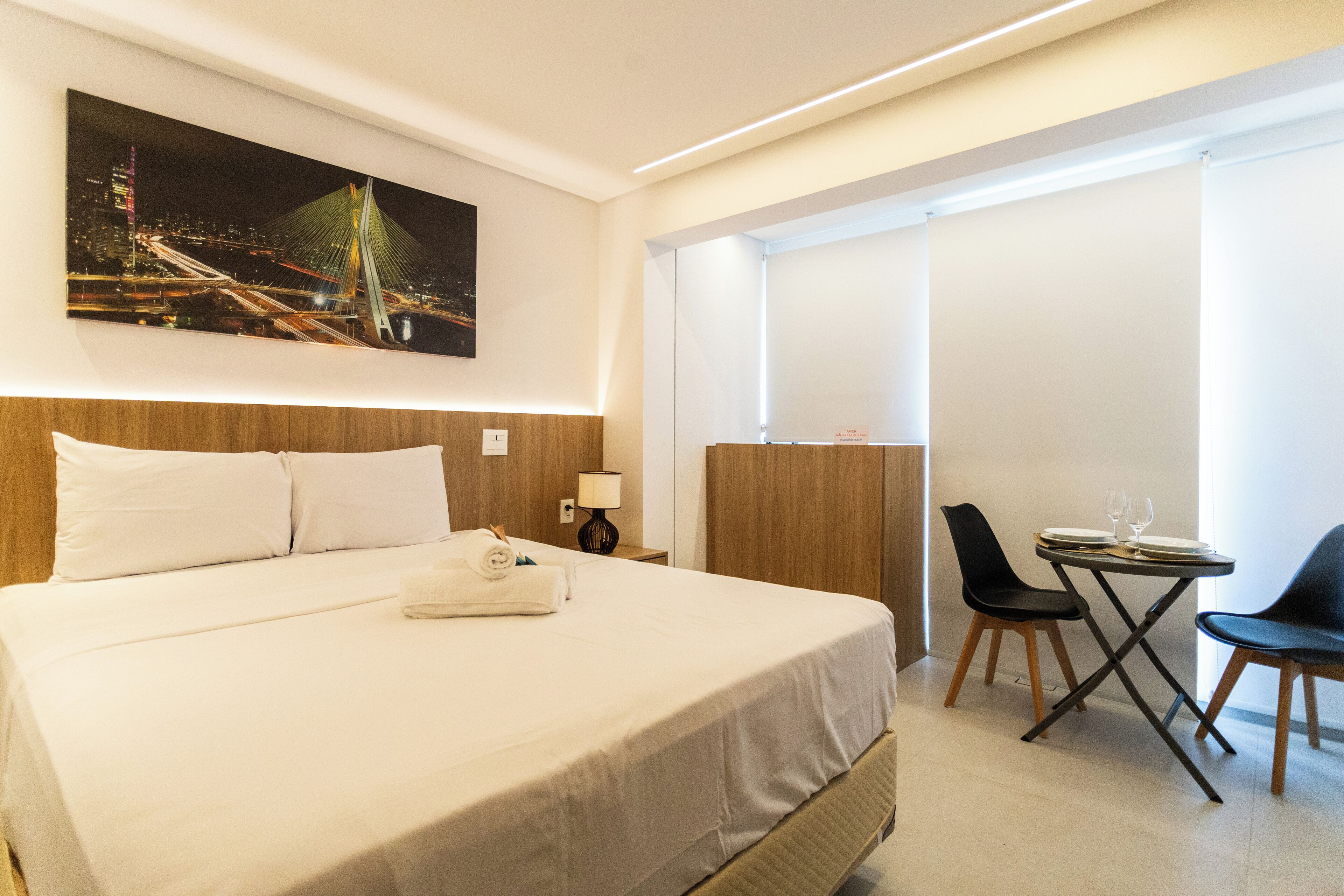 A hotel room with a large bed, a desk with two chairs, a painting of a bridge, and a window with blinds.