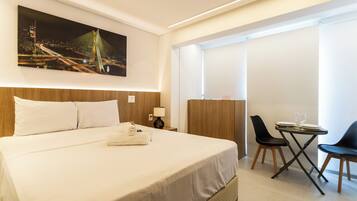 A hotel room with a large bed, a desk with two chairs, a painting of a bridge, and a window with blinds.