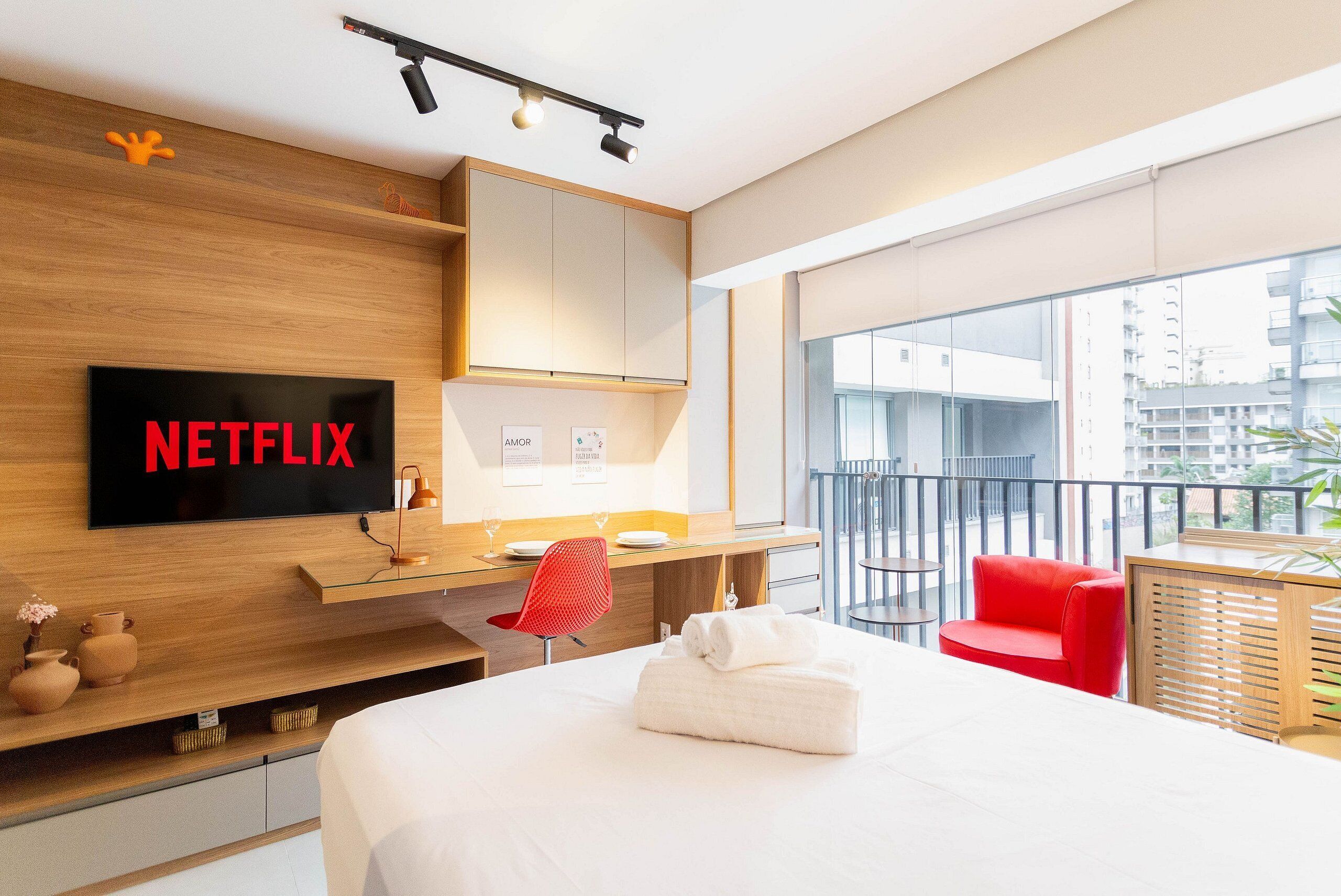 Ett modernt hotellrum med en stor säng, ett skrivbord med en platt-TV som visar ”NETFLIX”, en röd fåtölj och en balkong med utsikt över staden.