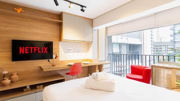 Ett modernt hotellrum med en stor säng, ett skrivbord med en platt-TV som visar ”NETFLIX”, en röd fåtölj och en balkong med utsikt över staden.