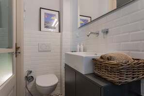 Une salle de bain avec des carreaux de céramique blancs rectangulaires, un lavabo blanc, un tableau encadré au mur et un panier en osier posé sur le comptoir.
