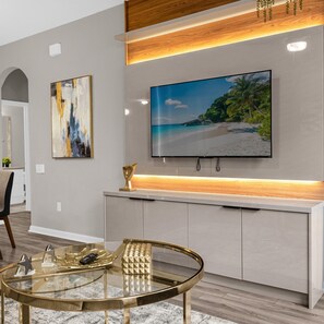 A modern living room with a large flat-screen TV mounted on a wooden shelf, a glass coffee table, and a minimalist design. - 2306 SP - Disney Villa 2BR Pool Near Magic (Kissimmee)
