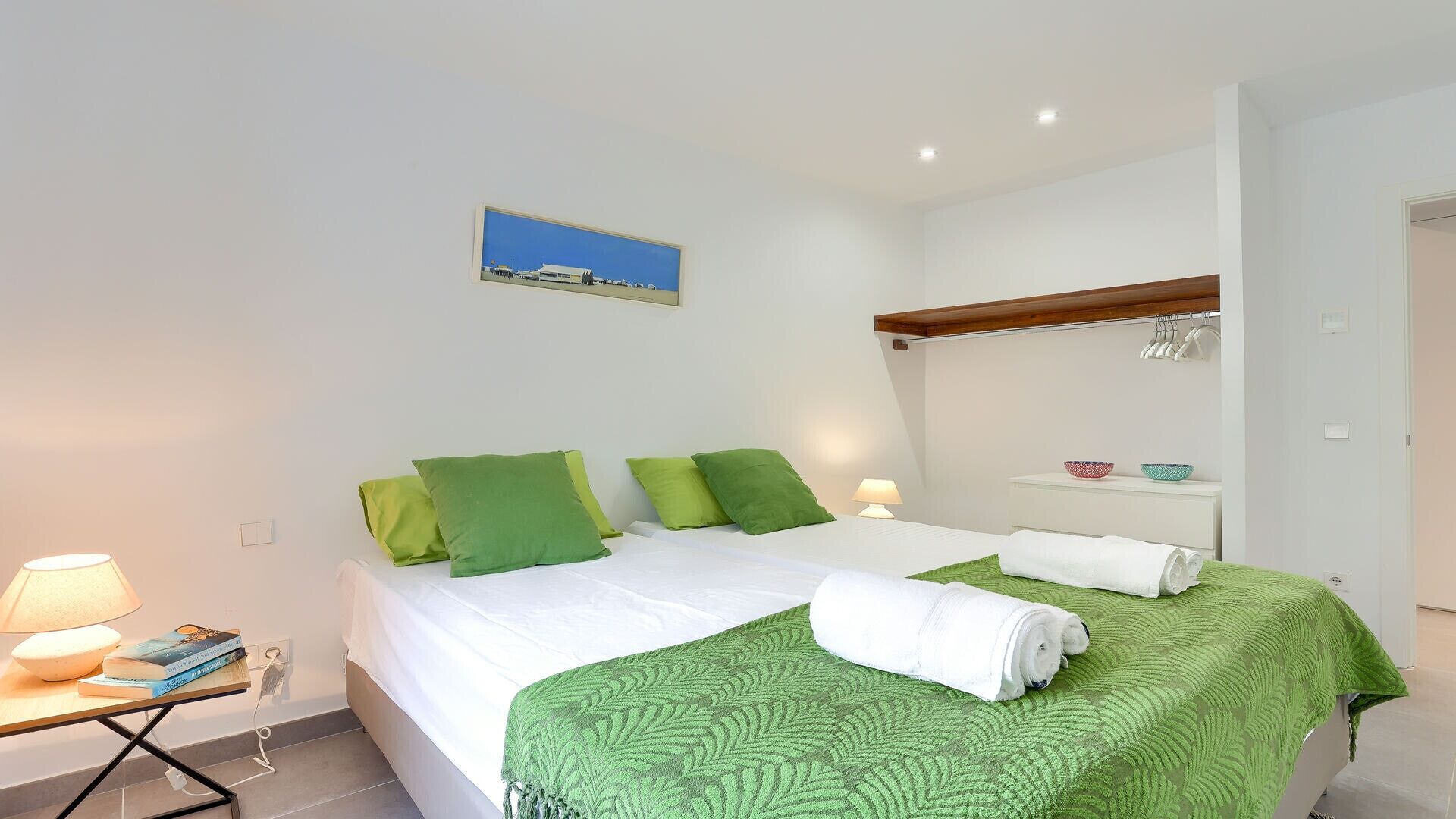 A hotel room with a bed, green pillows, a bedside table with a lamp, a small shelf with bowls, and a painting on the wall.