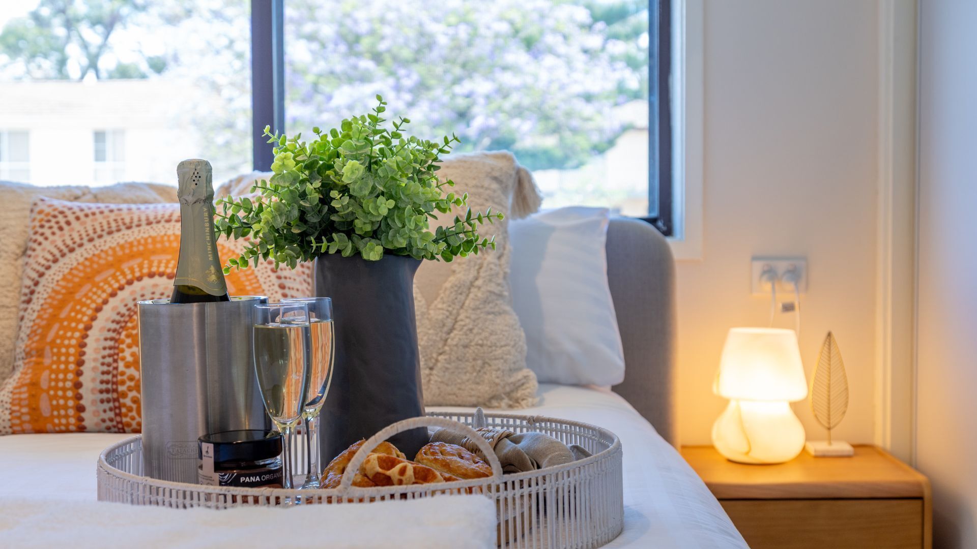 A cozy bedroom with a bed, a bedside table with a lamp, a window with a view of trees, a bedside plant, a tray with bread and a bottle, and a bottle of champagne.
