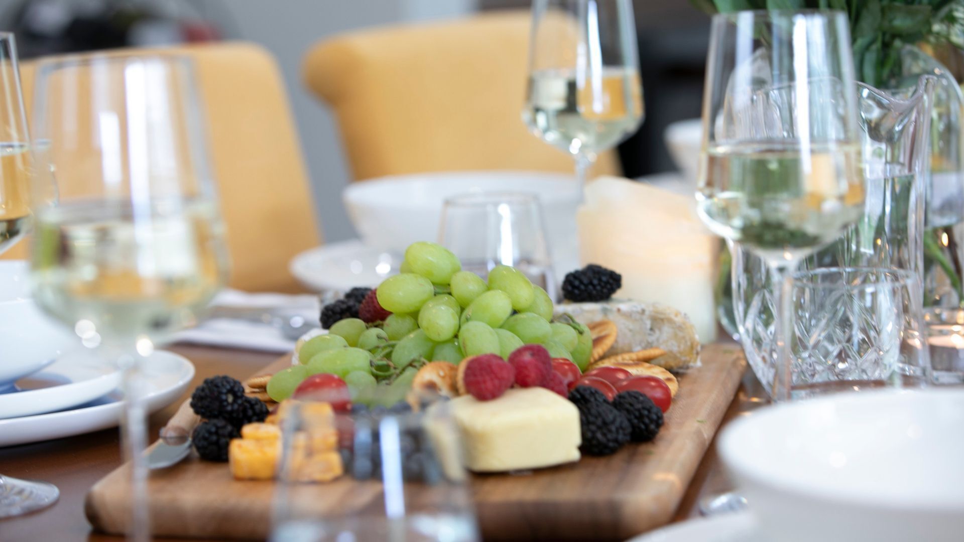 A table set for a meal with a fruit and cheese board, glasses of white wine, and candles.