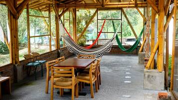 A covered outdoor dining area with a wooden table and chairs, surrounded by bamboo structures and large windows.