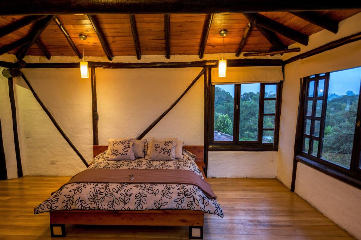Ein Schlafzimmer mit einem hölzernen Bett, Blick ins Freie durch große Fenster und einer geneigten Decke mit freiliegenden Balken.
