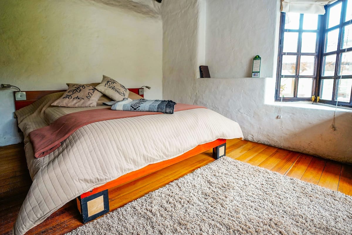 Ein Schlafzimmer mit einem Holzbettgestell, einer weißen Wand, einem Fenster mit Blick und einer kleinen Ablage mit einem grünen Schild.