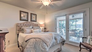 Une chambre à coucher avec une tête de lit capitonnée, un grand lit recouvert d’une literie à motifs, deux tableaux encadrés au mur, un ventilateur de plafond et une vue sur l’extérieur, visible à travers une porte-fenêtre coulissante.