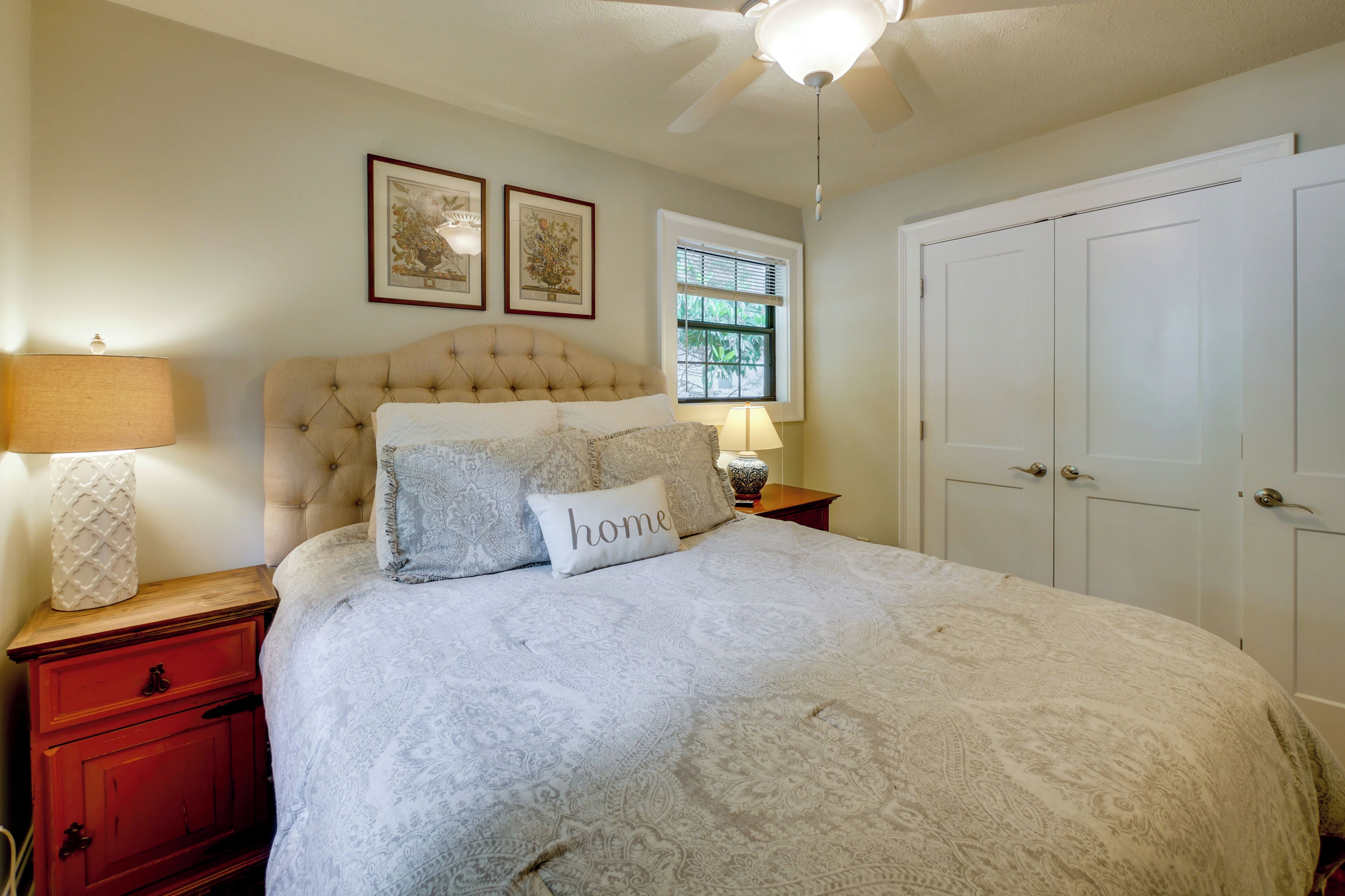 Une chambre à coucher avec une tête de lit capitonnée, un lit avec une literie à motifs, une table de chevet avec une lampe, une fenêtre avec des stores et une porte.