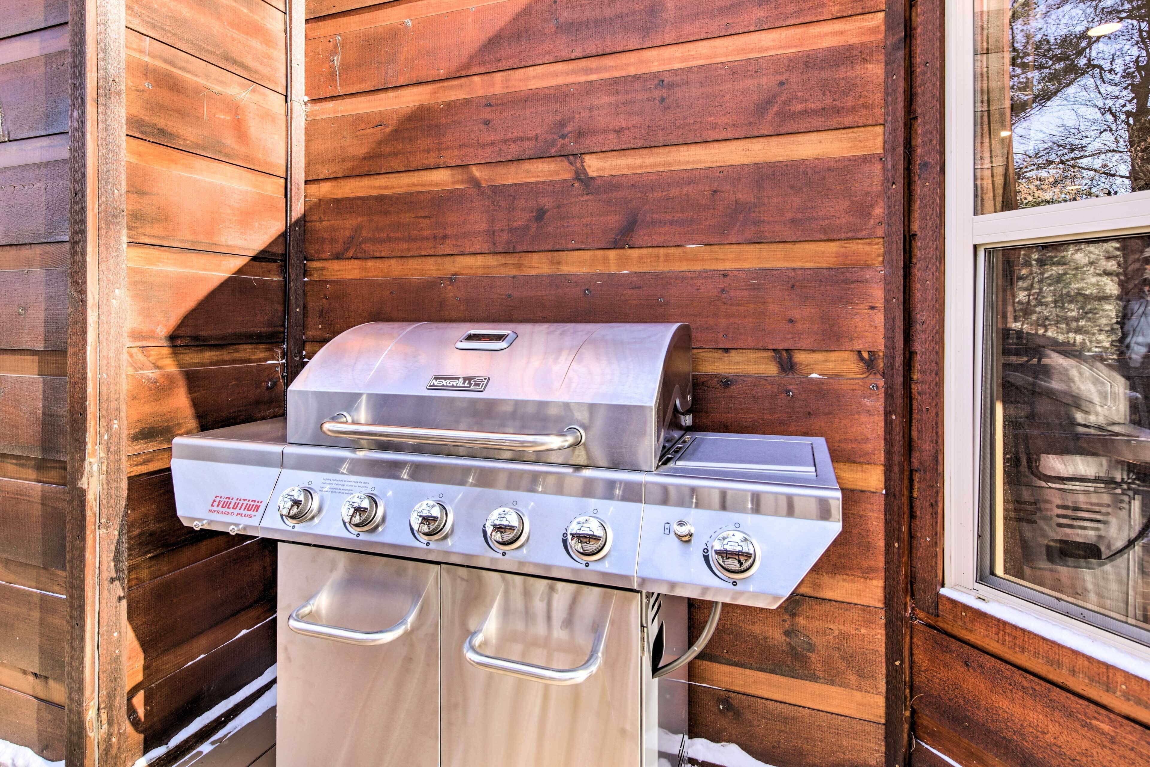 Un barbecue en acier inoxydable doté de quatre brûleurs, placé contre un mur en bois avec une fenêtre sur la droite.