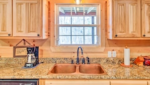 A kitchen with wooden cabinets, a granite countertop, a double sink, a coffee maker, and a window with a view of trees.