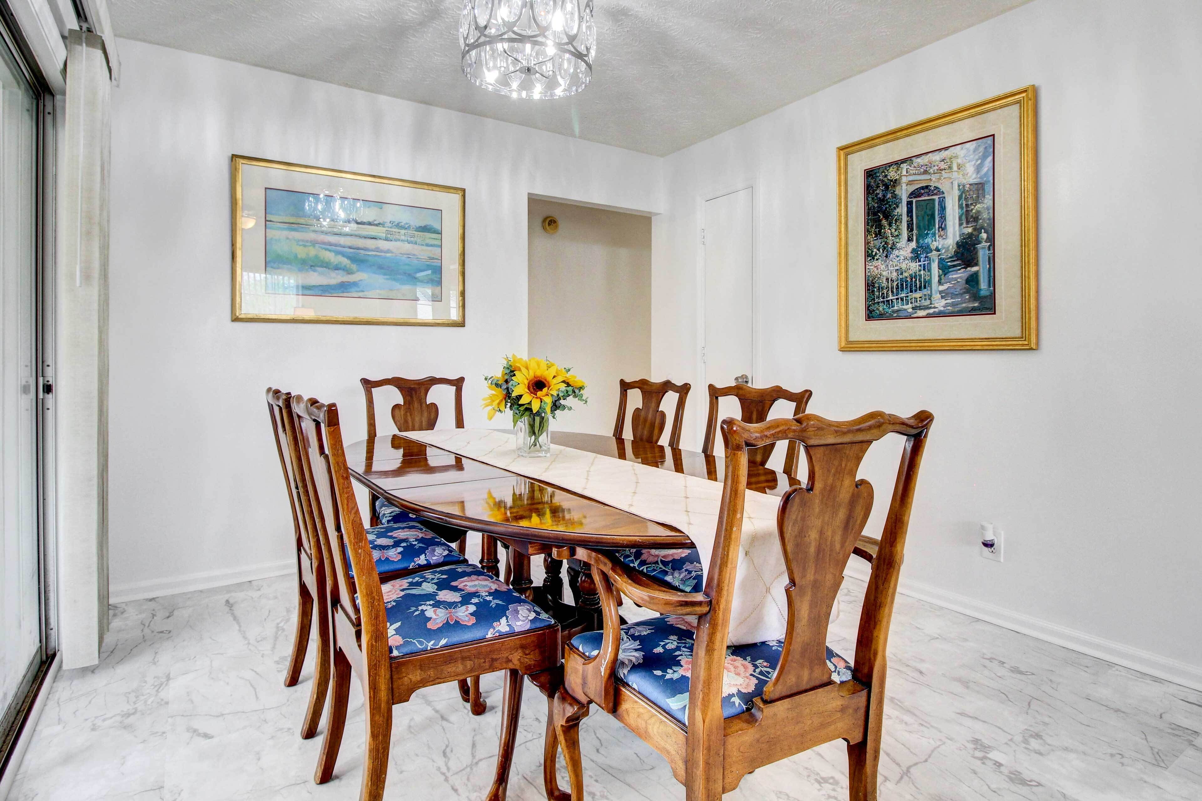 Uma sala de jantar com piso de mármore, cadeiras de madeira com estofamento floral, uma mesa de vidro e duas pinturas emolduradas nas paredes.