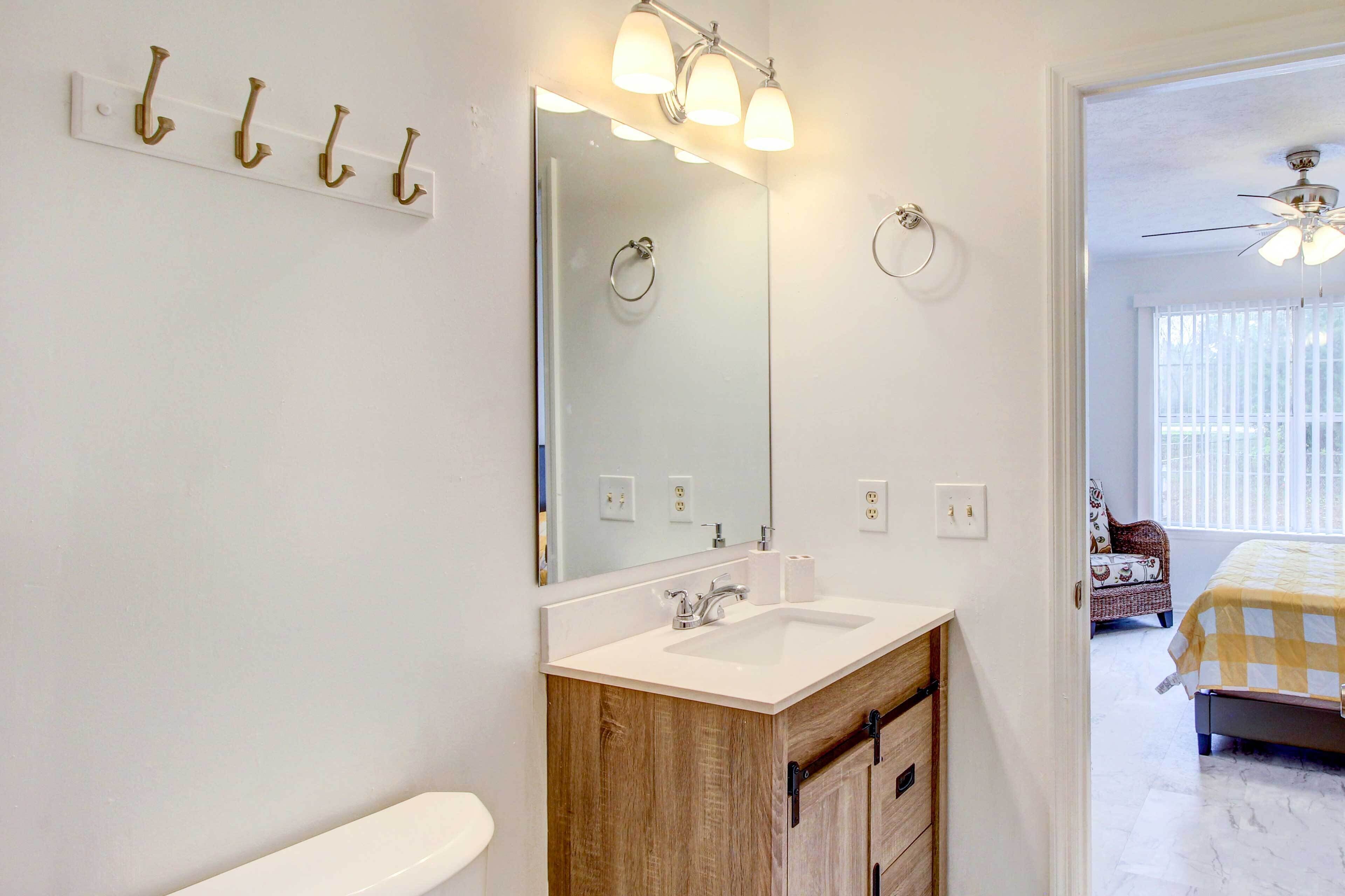 A bathroom with a white sink, wooden vanity, and a large mirror. There are hooks on the wall and a ceiling fan in the adjacent room.