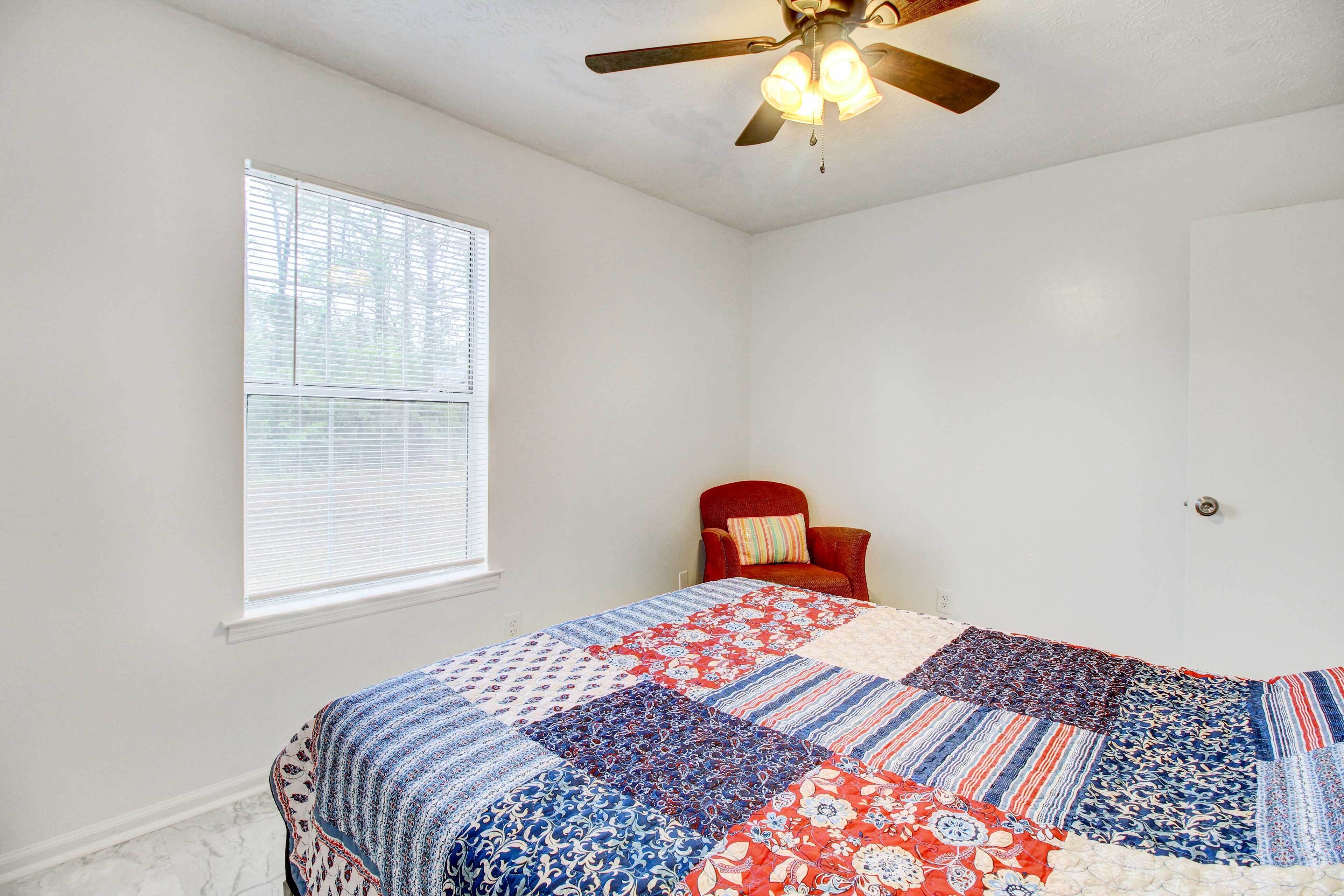 A bedroom with a large bed, a ceiling fan, a window with blinds, and a red chair.
