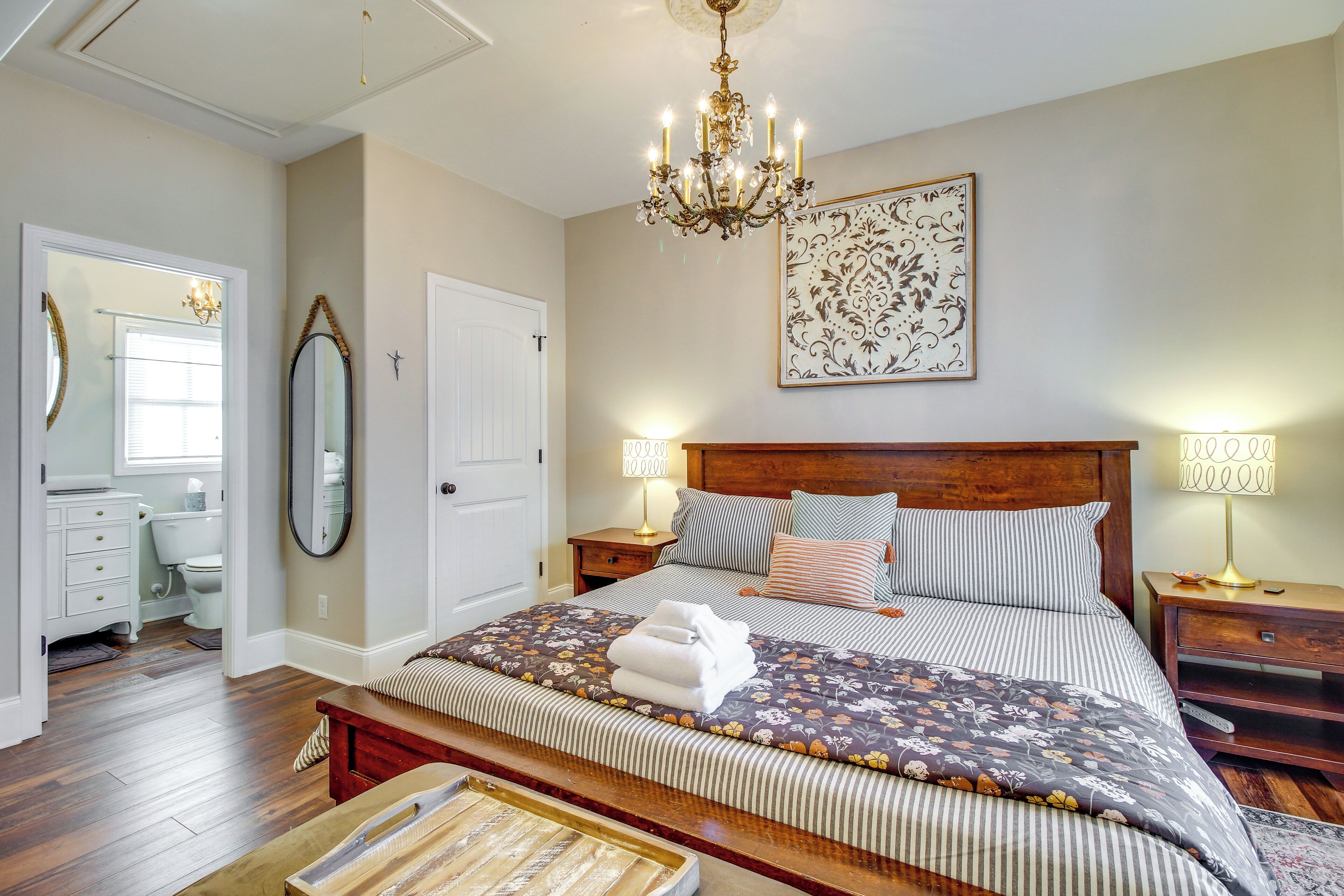 A bedroom with a wooden bed, two bedside tables, a chandelier, a large mirror, and a framed artwork on the wall.