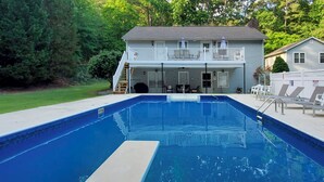 Une piscine rectangulaire dotée d’une plage blanche, entourée d’une pelouse et d’une maison avec un balcon.