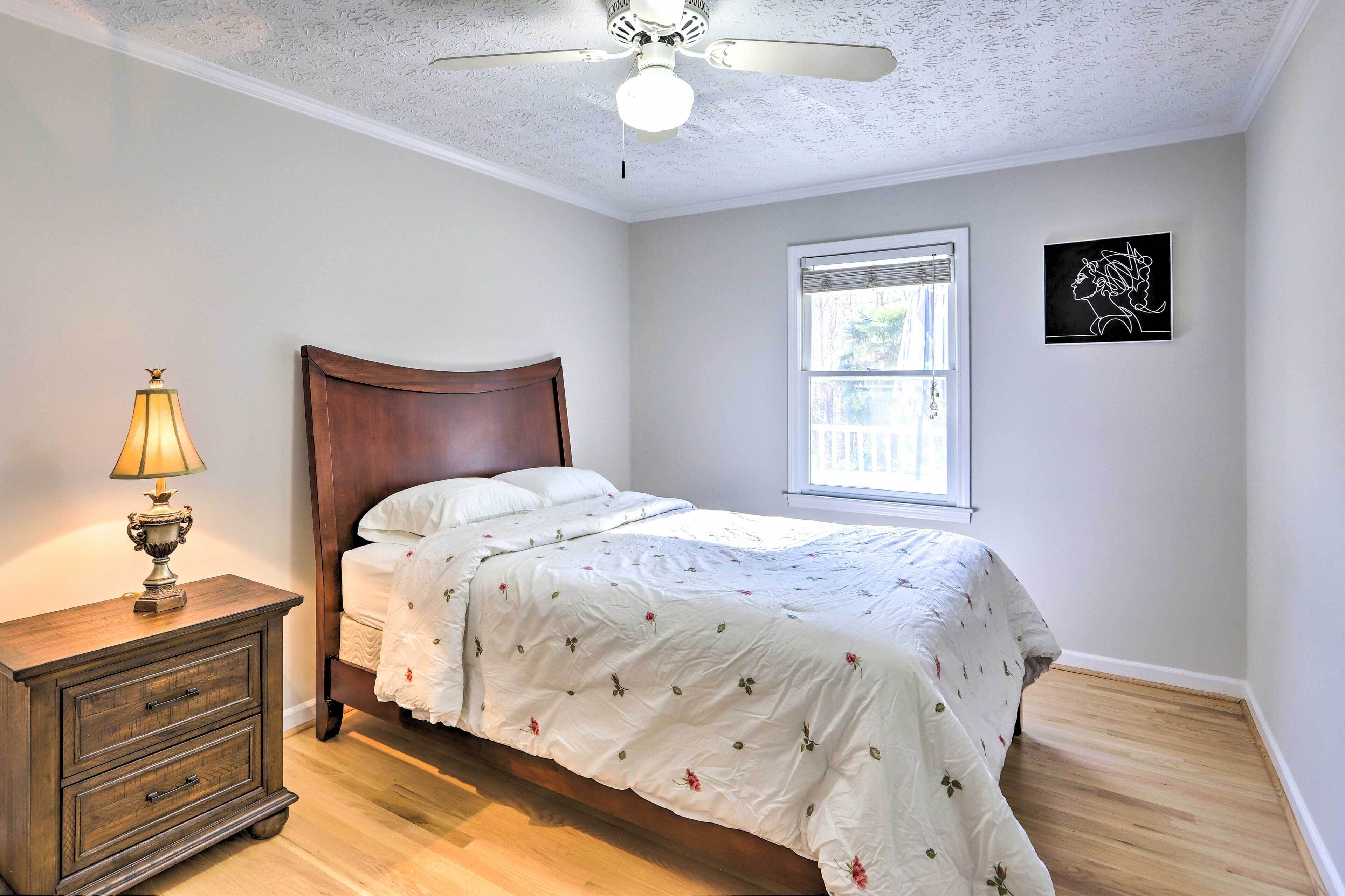 Un dormitorio con cama de madera, una mesita de noche con lámpara, ventilador de techo, ventana con persianas y una obra de arte en la pared.
