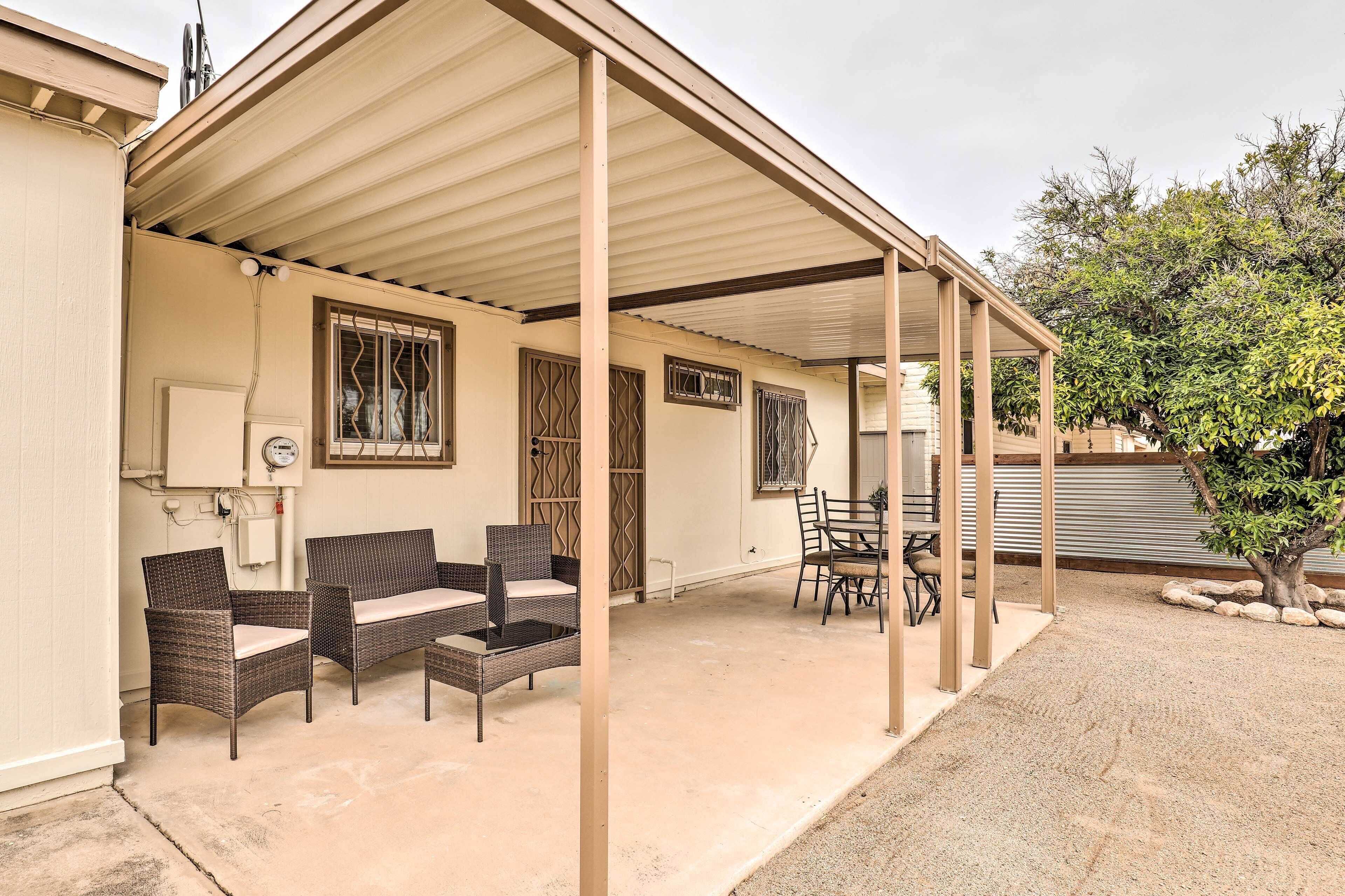A covered patio area with wicker furniture and a table, a window with decorative bars, and a tree in the background.