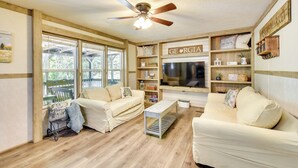 A cozy living room with two cream-colored sofas, a wooden coffee table, and a ceiling fan. A large window offers a view of greenery outside. Shelves along the walls hold decorative items and a sign that reads "GEORGIA."