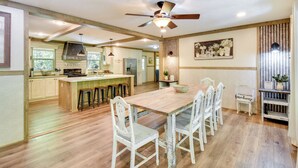 A spacious kitchen with wooden flooring, a central island with stools, a ceiling fan, and a dining area with white chairs.