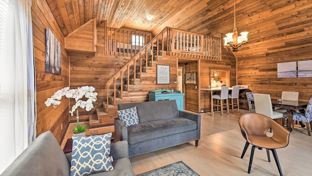 Interior de una cabaña de madera con sofá, sillón, mesa de comedor, sillas y una escalera.